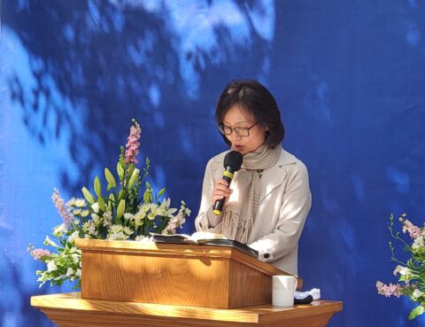 A woman speaking at a podium with floral decorations and blue background.