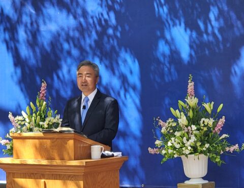 A man in a suit speaking at a wooden podium with floral arrangements.