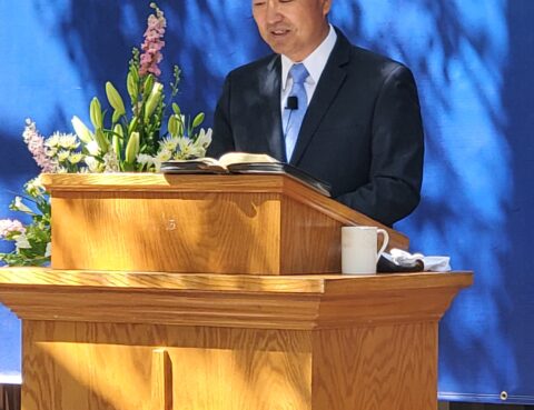 Man speaking at a wooden podium with flowers and a Bible.