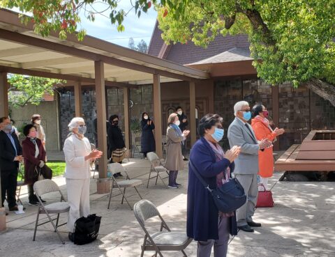 People wearing masks standing outside under a tree, observing social distancing.
