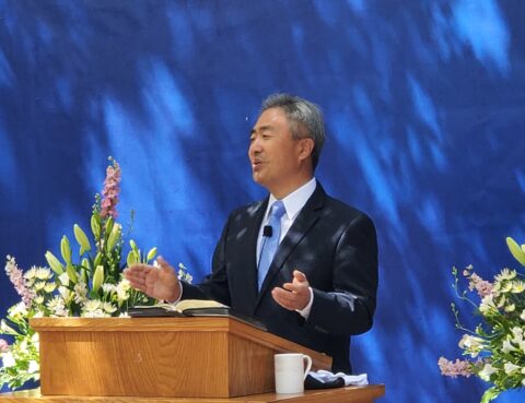 A man in a suit speaking at a podium with flowers and a blue background.