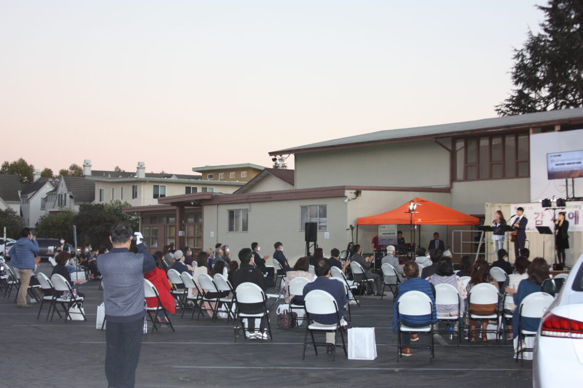 Outdoor gathering with people seated and a speaker addressing the crowd.