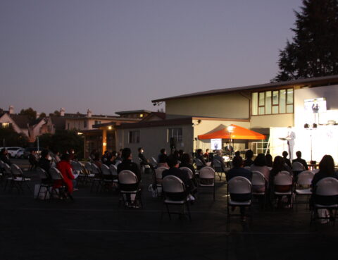 Outdoor evening event with an audience seated in front of a lit stage.
