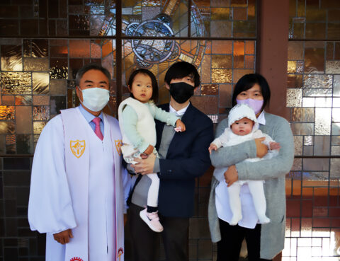 Family with a priest and two young children, all wearing masks indoors.