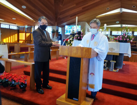 A clergyperson and a man in a church exchanging a handshake near a podium.