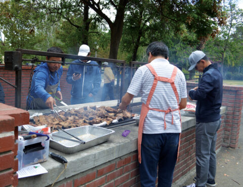 People grilling meat outdoors on a large barbecue grill.