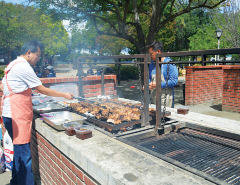 People grilling meat over an outdoor barbecue pit on a sunny day.