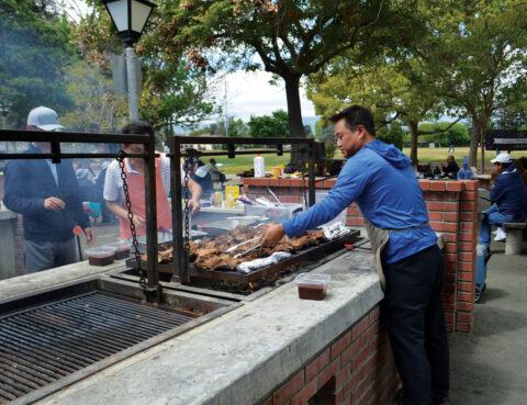 Man grilling meat outdoors at a large barbecue pit in a park.