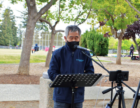 A man in a mask speaks outdoors at a microphone stand with a music sheet holder.