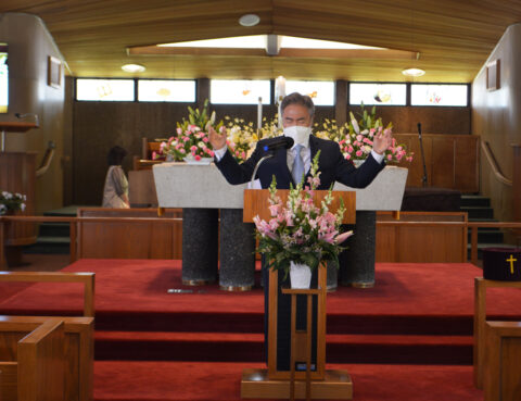 A pastor preaching inside a church with floral arrangements.