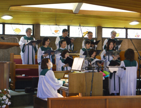 A choir performs with organ accompaniment in a sunlit room.