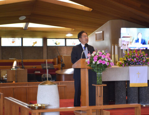 Man speaking at a wooden podium inside a church with floral arrangements.