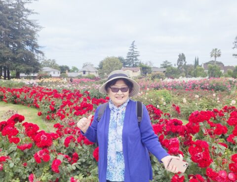 Smiling woman in purple jacket stands in vibrant red rose garden.