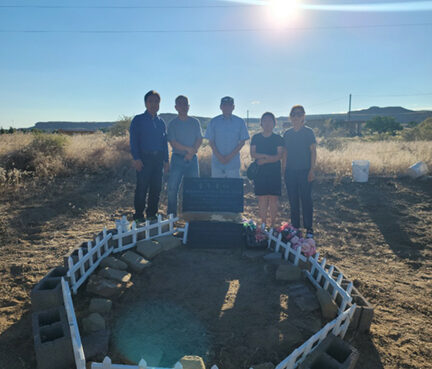 Four people standing around a small grave with a white picket fence in a sunny, open field.