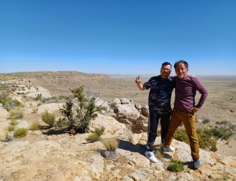 Two men smiling and posing in a desert landscape under a clear blue sky.