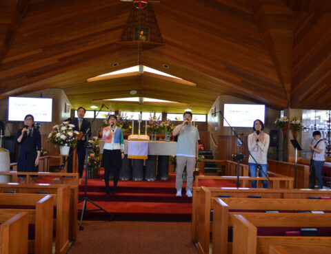 Four people standing on a church stage, wearing masks and holding papers.
