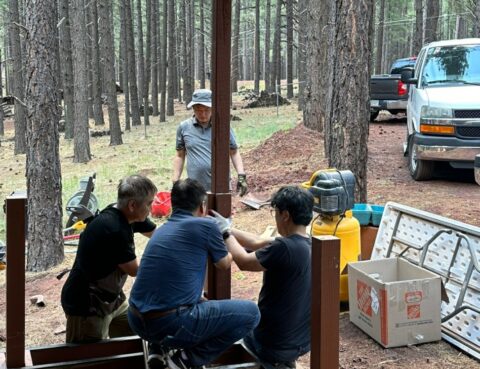 People gathered outdoors in a wooded area near a parked vehicle and supplies.