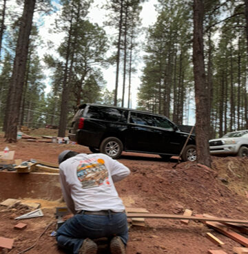 A person kneeling on a dirt path near a parked black SUV in a forest.