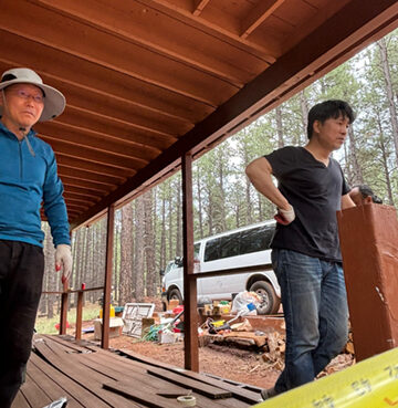 Two men standing on a wooden deck surrounded by camping gear and trees.