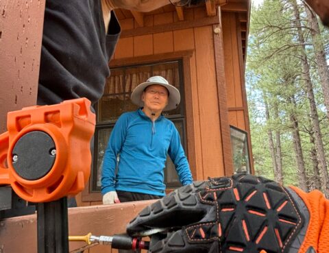 Two people outside a wooden cabin with tools and gloves in the foreground.