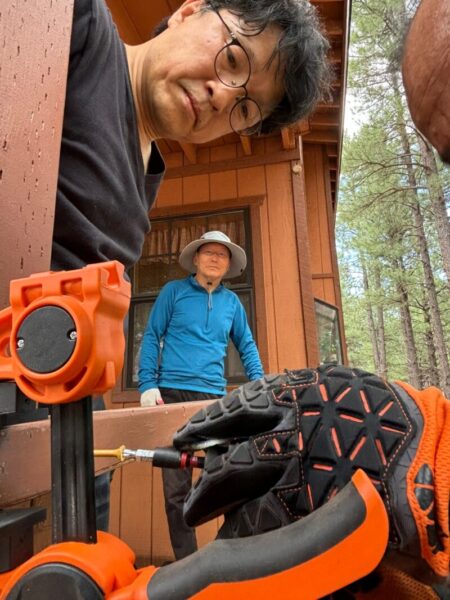 Two people outside a wooden cabin with tools and gloves in the foreground.