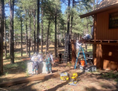 Group renovating a wooden house in a forested area with tools and ladders.